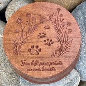 May include: A round, wooden keepsake box with a carved design. The lid features paw prints, flowers, and the inscription "You left paw prints on our hearts." The box is a warm brown color and sits on a bed of stones.