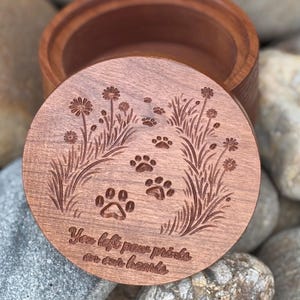 May include: A wooden keepsake box with a lid featuring engraved paw prints, flowers, and grass. The lid has the text "You left paw prints on our hearts." The box is made of a light brown wood.