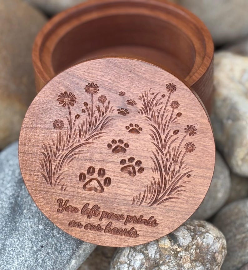 May include: A wooden keepsake box with a lid featuring engraved paw prints, flowers, and grass. The lid has the text "You left paw prints on our hearts." The box is made of a light brown wood.