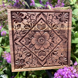 May include: Square wooden coaster with intricate floral carvings. The coaster features a central diamond-shaped design with a large flower and surrounding floral and leaf patterns. The wood has a warm, natural tone.