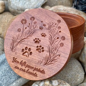May include: A round, wooden box with a lid. The lid is engraved with paw prints, flowers, and the text "You left paw prints on our hearts." The box is a warm brown color and sits on a bed of rocks.