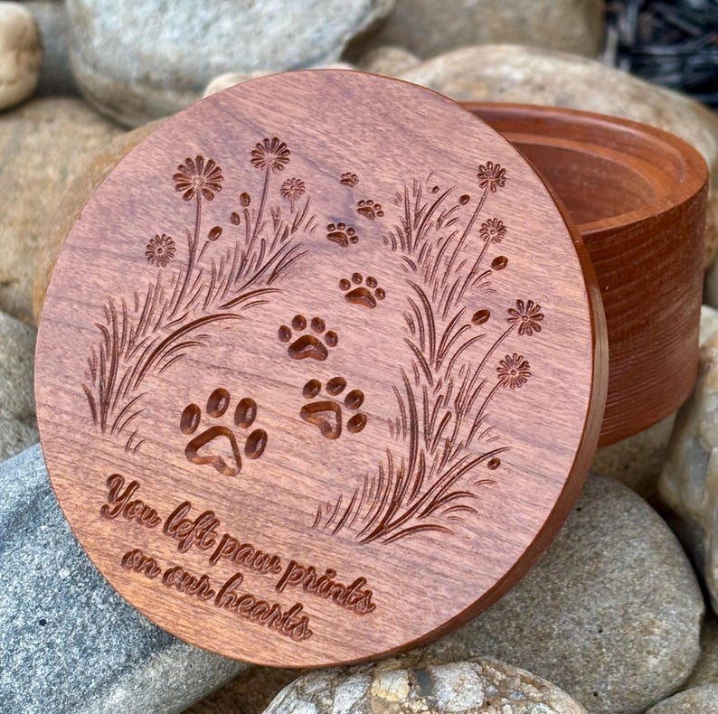 May include: A round, wooden box with a lid. The lid is engraved with paw prints, flowers, and the text "You left paw prints on our hearts." The box is a warm brown color and sits on a bed of rocks.