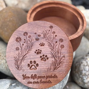 May include: A wooden pet memorial box with a lid featuring engraved paw prints and floral designs. The lid has the inscription "You left paw prints on our hearts." The box is made of a reddish-brown wood.