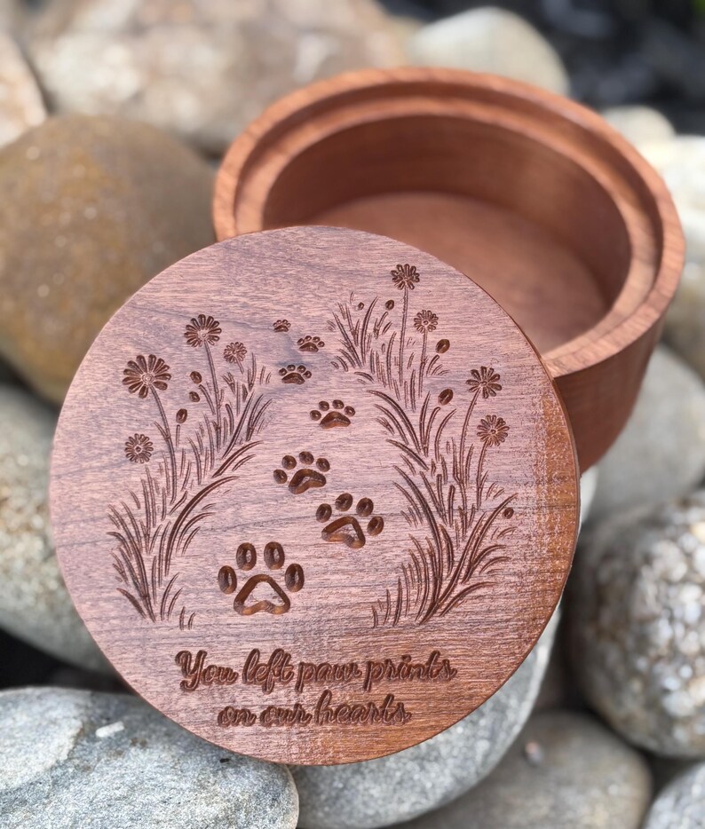 May include: A wooden pet memorial box with a lid featuring engraved paw prints and floral designs. The lid has the inscription "You left paw prints on our hearts." The box is made of a reddish-brown wood.