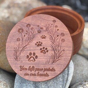 May include: A wooden keepsake box with a lid featuring engraved paw prints and floral designs. The lid has the text "You left paw prints on our hearts". The box is made of reddish-brown wood.