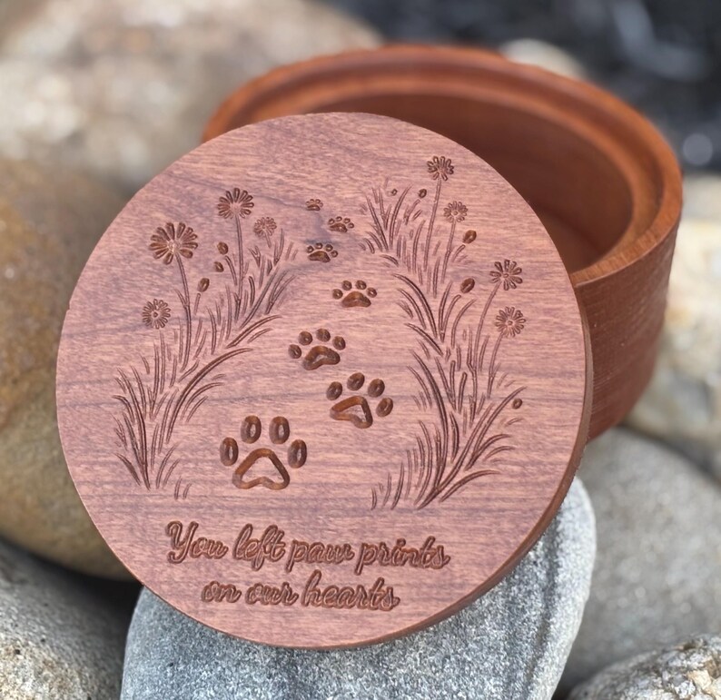 May include: A wooden keepsake box with a lid featuring engraved paw prints and floral designs. The lid has the text "You left paw prints on our hearts". The box is made of reddish-brown wood.