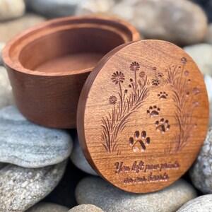 May include: A wooden pet urn with a lid. The lid features engraved paw prints, flowers, and the text "You left paw prints on our hearts." The urn is open, revealing its interior. The wood has a warm, reddish-brown tone.
