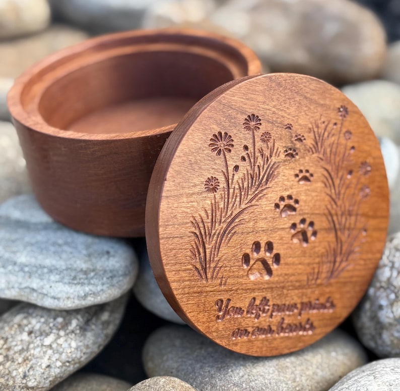 May include: A wooden pet urn with a lid. The lid features engraved paw prints, flowers, and the text "You left paw prints on our hearts." The urn is open, revealing its interior. The wood has a warm, reddish-brown tone.