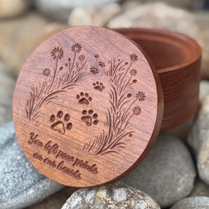 May include: A round, wooden keepsake box with a lid featuring engraved paw prints and floral designs. The lid has the inscription "You left paw prints on our hearts." The box is made of reddish-brown wood.
