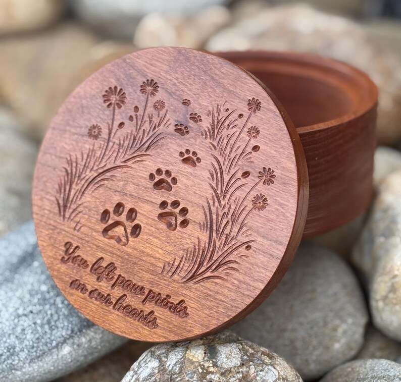May include: A round, wooden keepsake box with a lid featuring engraved paw prints and floral designs. The lid has the inscription "You left paw prints on our hearts." The box is made of reddish-brown wood.