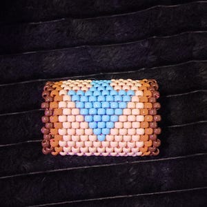 May include: A rectangular beaded bracelet with a geometric design. The bracelet is mainly tan, with a blue heart shape in the centre. The edges are brown, and the beads are arranged in a grid pattern.