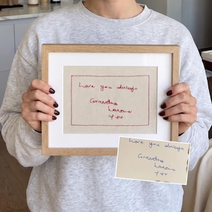 Op de afbeelding: Een lichthouten lijst met een beige stoffen paneel met rood geborduurde tekst die luidt Love you always Grandma Larane y y. Een kleine inzet toont het originele handgeschreven briefje in blauwe inkt op wit papier met dezelfde tekst.