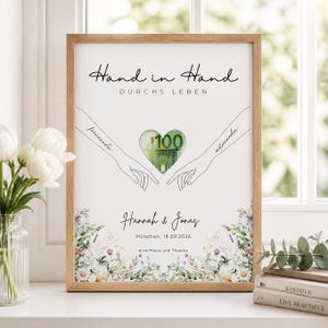 Può includere: Una stampa incorniciata con il testo Hand in Hand DURCHS LEBEN, due braccia disegnate a tratto verso un ritaglio a forma di cuore che rivela una banconota da 100 euro, personalizzata con Hannah & Jonas, München, 18.09.2026, e illustrazioni floreali.