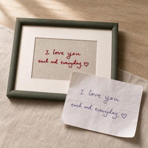 Op de afbeelding: Een donkergroene rechthoekige fotolijst met een beige stoffen inzetstuk met de in rood geborduurde tekst "I love you each and everyday." en een klein hartje. Een wit vel papier met dezelfde tekst in blauwe inkt ligt ervoor.