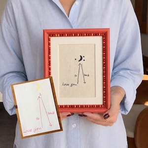Könnte beinhalten: Ein rechteckiger Bilderrahmen mit rotem Perlenrand enthält einen cremefarbenen Stoffeinsatz mit schwarzer Stickerei "Love you to the moon and back" sowie einem Halbmond- und Punktemotiv, dazu eine passende Karte.