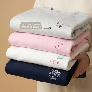 Könnte beinhalten: Ein Stapel gefalteter Sweatshirts in Grau, Rosa, Weiß und Marineblau. Jedes Sweatshirt hat eine individuelle Stickerei, darunter Namen, Sätze wie "Best Mom Ever" und familienbezogene Grafiken. Das oberste Sweatshirt hat den Text "with your own personal design."
