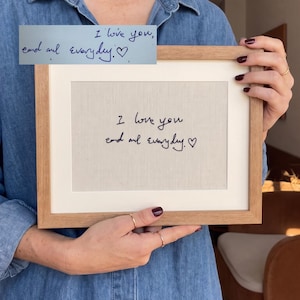Può includere: Una cornice rettangolare in legno contenente un inserto in tessuto beige chiaro con il testo nero ricamato "I love you and everyday." seguito da un simbolo a forma di cuore. Il ricamo replica lo stile manoscritto dell'immagine inserita.