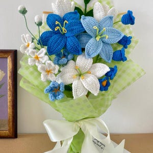 Peut inclure: Un bouquet de fleurs au crochet composé de lys dans des tons bleu roi, bleu clair et blanc, agrémenté de petits boutons blancs et de fleurs bleues en forme de cloche. L'ensemble est enveloppé dans du papier à carreaux vert clair et noué d'un ruban blanc.