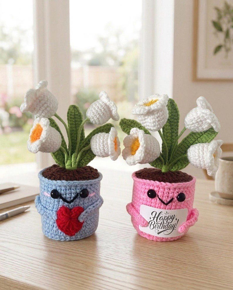 May include: Two crocheted potted plants with white flowers and green leaves. The blue pot features a red heart, while the pink pot holds a white sign with the text Happy Birthday. Both pots have black bead eyes and embroidered smiling mouths.