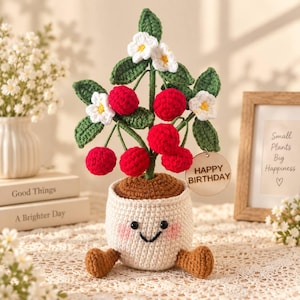 May include: A crocheted cherry tree in a cream-coloured pot with a smiling face, pink cheeks, and brown feet. The tree features green leaves, white flowers with yellow centres, and bright red cherries. A round wooden tag reads HAPPY BIRTHDAY.