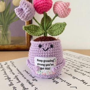 Könnte beinhalten: Ein handgehäkelter Blumentopf mit drei Tulpen in Rosa- und Lilatönen. Der lavendelfarbene Topf hat ein lächelndes Gesicht und ein Schild mit der Aufschrift "Keep growing strong you've got this!". Ein bezauberndes Deko-Objekt.