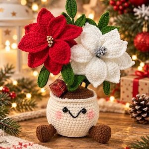 May include: A crocheted amigurumi plant in a cream-coloured pot with a smiling face, pink cheeks, and brown feet. The plant features one large red poinsettia with gold centres and one white poinsettia with silver centres, surrounded by green leaves.