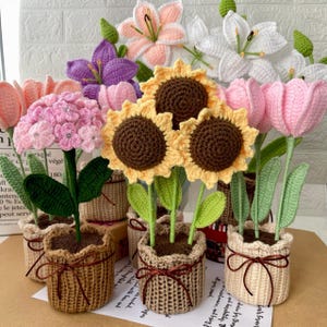 Peut inclure: Une collection de fleurs au crochet faites à la main, dont des tournesols, des lys et des tulipes, dans de petits paniers tressés. Les fleurs sont de différentes couleurs, comme le jaune, le rose, le violet et le blanc. Chaque fleur a des feuilles et des tiges vertes.