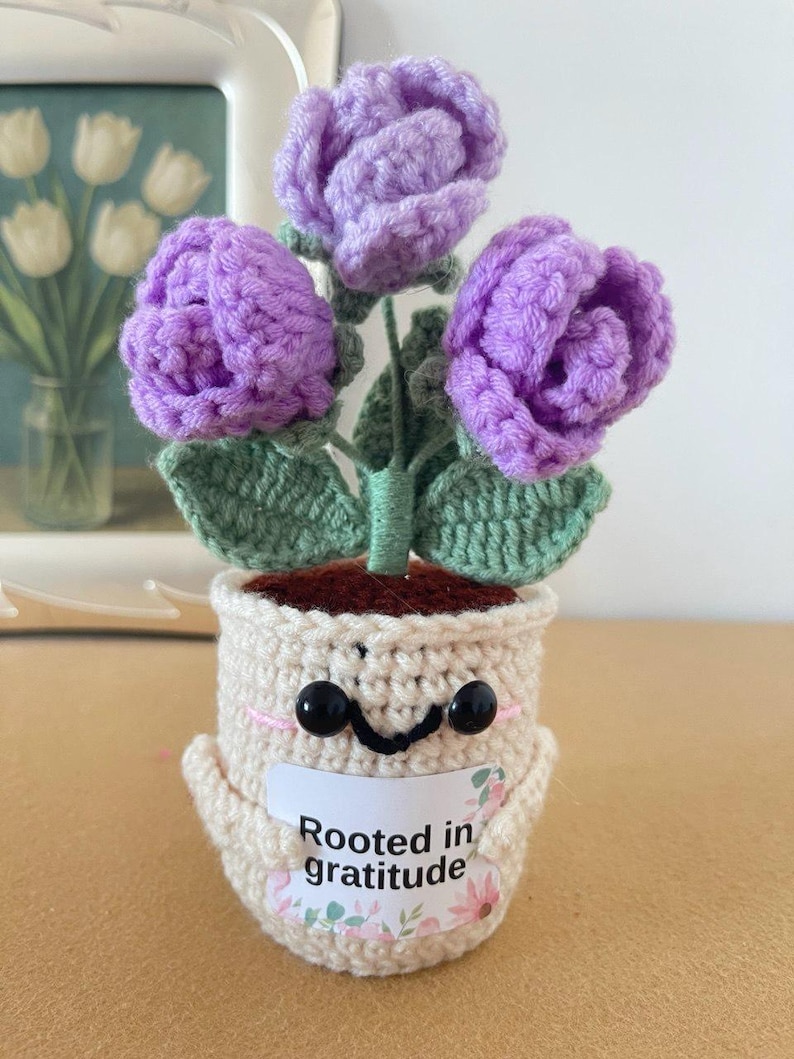 May include: A crocheted potted plant featuring three purple flowers with green leaves. The cream-colored pot has a smiling face with black bead eyes and holds a white sign that reads Rooted in gratitude with small pink and green floral accents.