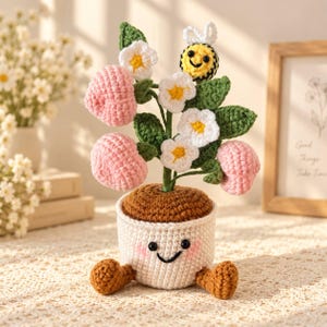 May include: A crocheted potted plant featuring a white pot with a smiling face, pink cheeks, and brown feet. The plant has green leaves, white flowers with yellow centers, pink rounded fruit shapes, and a small yellow and black bee with white wings.