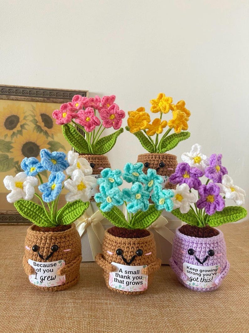 May include: Five crocheted potted flowers with smiling faces and small signs. The pots are brown or light purple, holding pink, yellow, blue, white, turquoise, and purple flowers with green leaves. The signs feature text including Because of you I grew, A small thank you that grows, and Keep growing strong you've got this!