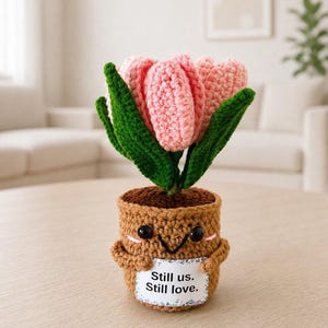 May include: A crocheted pink tulip plant in a brown pot with a smiling face. The pot features small arms holding a white rectangular sign that reads "Still us. Still love." The tulip has textured light pink petals and dark green leaves.