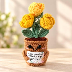 Op de afbeelding: Een gehaakte potplant met drie felgele bloemen en groene bladeren. De bruine pot heeft een lachend gezicht met zwarte ogen en houdt een wit bordje vast met de tekst Keep growing strong you've got this! en bloemmotieven.