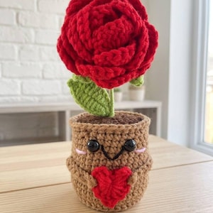 May include: A crocheted red rose with a green leaf sits in a tan crocheted pot. The pot features a smiling face with black bead eyes, a stitched mouth, and pink cheeks, while holding a small red crocheted heart against its front surface.