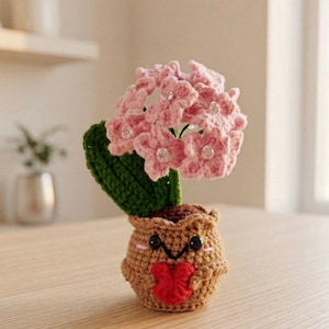 May include: A crocheted potted plant featuring a light brown pot with a smiling face and a red heart held in its small arms. The plant has a single dark green leaf and a cluster of light pink flowers, each accented with a small clear bead at the center.
