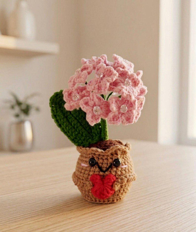 May include: A crocheted potted plant featuring a light brown pot with a smiling face and a red heart held in its small arms. The plant has a single dark green leaf and a cluster of light pink flowers, each accented with a small clear bead at the center.
