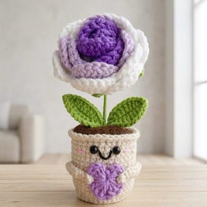 May include: A crocheted flower in a cream-colored pot with a smiling face, holding a small purple heart. The flower has a deep purple center, light purple middle petals, and white outer petals, with two green leaves on a thin green stem.
