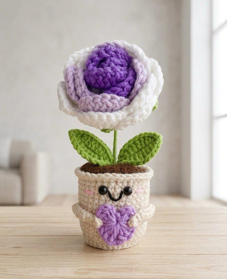 May include: A crocheted flower in a cream-colored pot with a smiling face, holding a small purple heart. The flower has a deep purple center, light purple middle petals, and white outer petals, with two green leaves on a thin green stem.