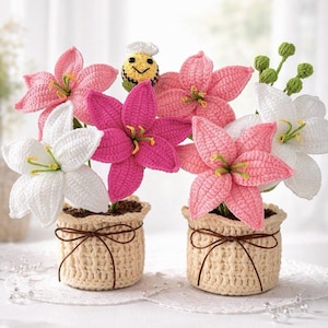 Peut inclure: Deux arrangements floraux au crochet dans des pots beiges. Les fleurs sont roses et blanches, avec des centres jaunes et des accents verts. Une petite figurine d'abeille souriante est nichée parmi les fleurs. Les pots sont attachés avec de la ficelle brune.