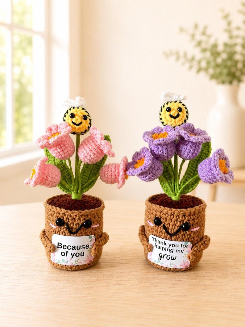 May include: Two crocheted potted plants with smiling faces. One pot holds pink flowers and a bee, with a sign reading "Because of you". The other pot holds purple flowers and a bee, with a sign reading "Thank you for helping me grow".