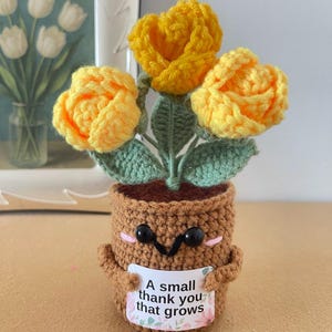 May include: A crocheted brown pot with a smiling face, black eyes, and pink cheeks, holding a white sign that reads A small thank you that grows. The pot contains three bright yellow crocheted flowers with green leaves and stems.
