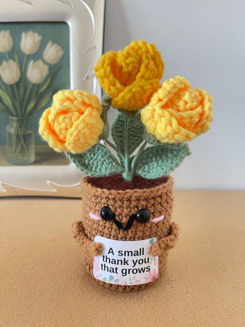 May include: A crocheted brown pot with a smiling face, black eyes, and pink cheeks, holding a white sign that reads A small thank you that grows. The pot contains three bright yellow crocheted flowers with green leaves and stems.