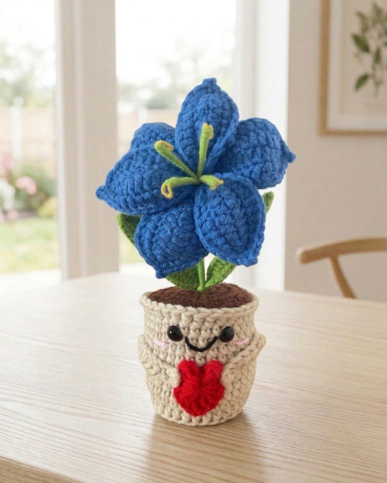 May include: A crocheted blue lily flower with green stems and leaves planted in a beige crocheted pot. The pot features a smiling face with black eyes, a black mouth, pink cheeks, and two small arms holding a red crocheted heart shape.