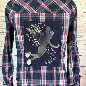 May include: A long-sleeved plaid shirt in navy blue, pink, and white. The back features a navy blue patch with a silver fairy design and scattered sequins. The shirt has buttoned cuffs and a classic collar.