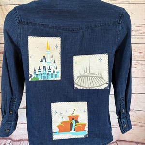May include: Dark blue denim shirt with three rectangular patches featuring colorful illustrations. The patches depict a castle, a futuristic structure, and a landscape with waterfalls. The shirt has buttoned cuffs and a collar.