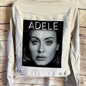 May include: A light beige denim jacket featuring a black and white portrait of Adele on the back. The portrait is framed by a black rectangle with the name "ADELE" in white block letters above the image. The jacket has a button-down front and two chest pockets.