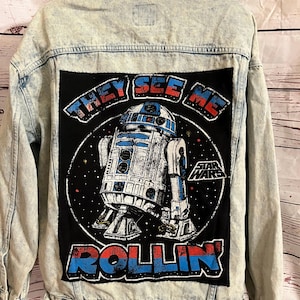 May include: Acid wash denim jacket with a large graphic on the back featuring R2-D2 from Star Wars. The graphic reads "THEY SEE ME ROLLIN'" in blue and red lettering. The jacket has a button-up front and two chest pockets.