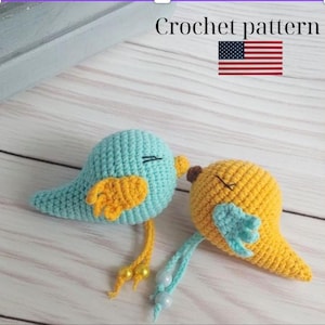 May include: Two crocheted bird ornaments, one blue and one yellow, with orange wings and beaded accents. The birds have closed eyes, suggesting sleep or rest. The text "Crochet pattern" and an American flag are in the background.