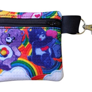 May include: A small, square coin purse featuring a colorful Care Bears design. The purse has a black zipper, a black strap, and a silver-colored clasp. The design includes purple and blue bears, rainbows, and clouds.