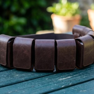 May include: A dark brown utility belt featuring multiple rectangular pouches with flap closures. The belt is constructed from a smooth, textured material and is designed to wrap around the waist, providing several storage compartments for small items.