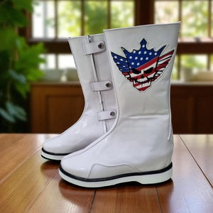 May include: Pair of white, high-top boots with a glossy finish. Each boot features a black sole and a decorative patch with a skull, crown, and American flag design. The boots have buckles and a zipper closure.